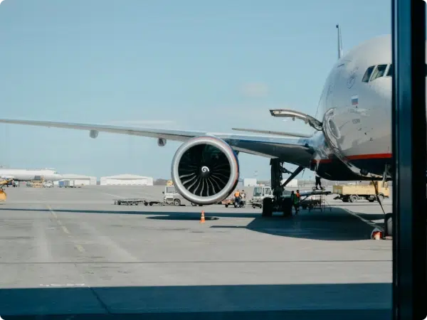 Airplane engine close-up with baggage carts and hangars visible on the tarmac under a clear blue sky.