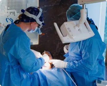 Two dental professionals in blue scrubs and masks performing a procedure on a patient in a clinical setting.